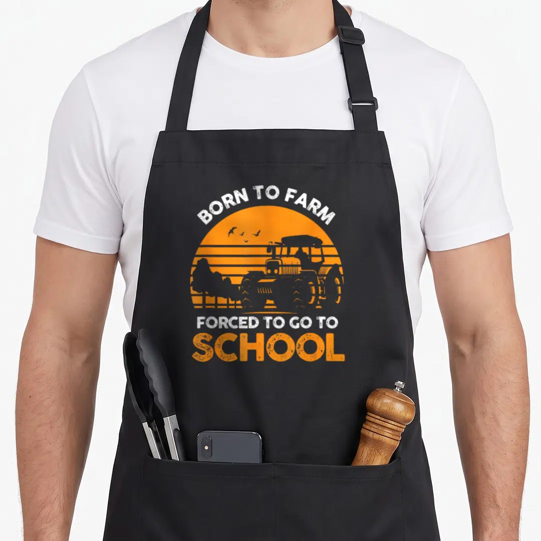 School farmer apron - Retro orange sunset tractor graphic with born to farm forced to go to school text — shown on a black cotton canvas apron against a white background