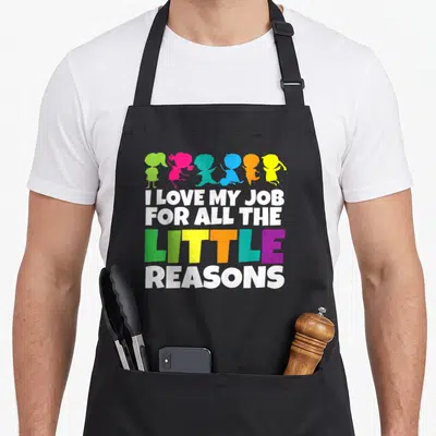 Retro reason teacher apron - I Love My Job For All The Little Reasons text with six colorful child silhouettes in green — shown on a black cotton canvas apron against a white background