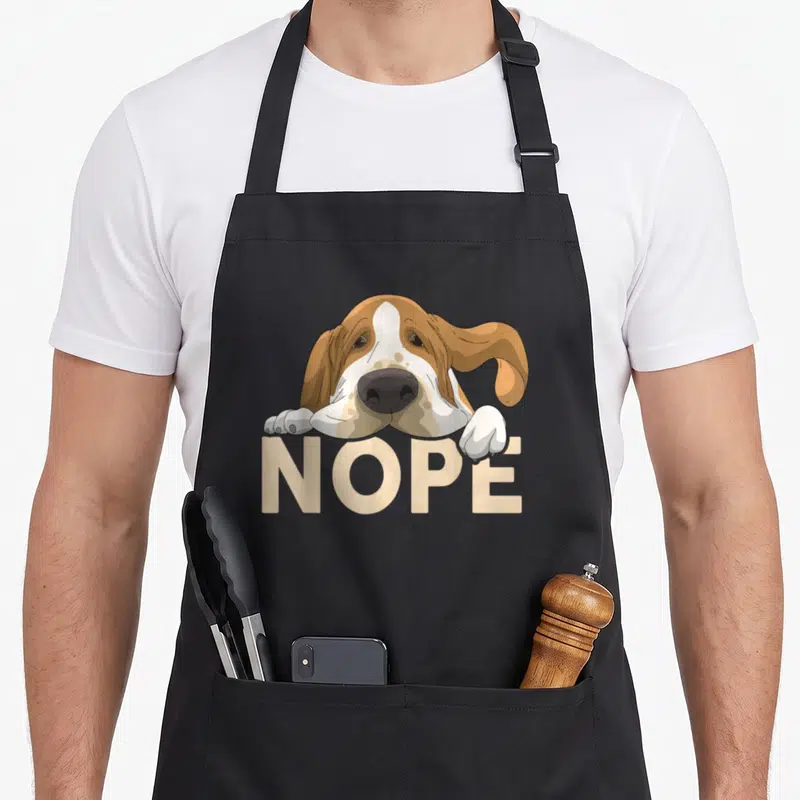 Hound nope dog apron - Cartoon basset hound resting paws on beige NOPE text — shown on a black cotton canvas apron against a white background