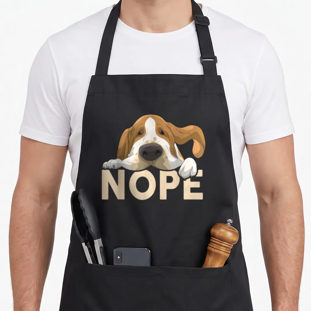 Hound nope dog apron - Cartoon basset hound resting paws on beige NOPE text — shown on a black cotton canvas apron against a white background