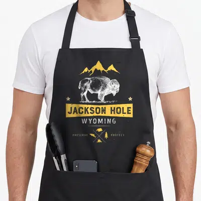 Jackson buffalo apron - White bison under yellow mountains with Jackson Hole text and diamond camping icons — shown on a black cotton canvas apron against a white background
