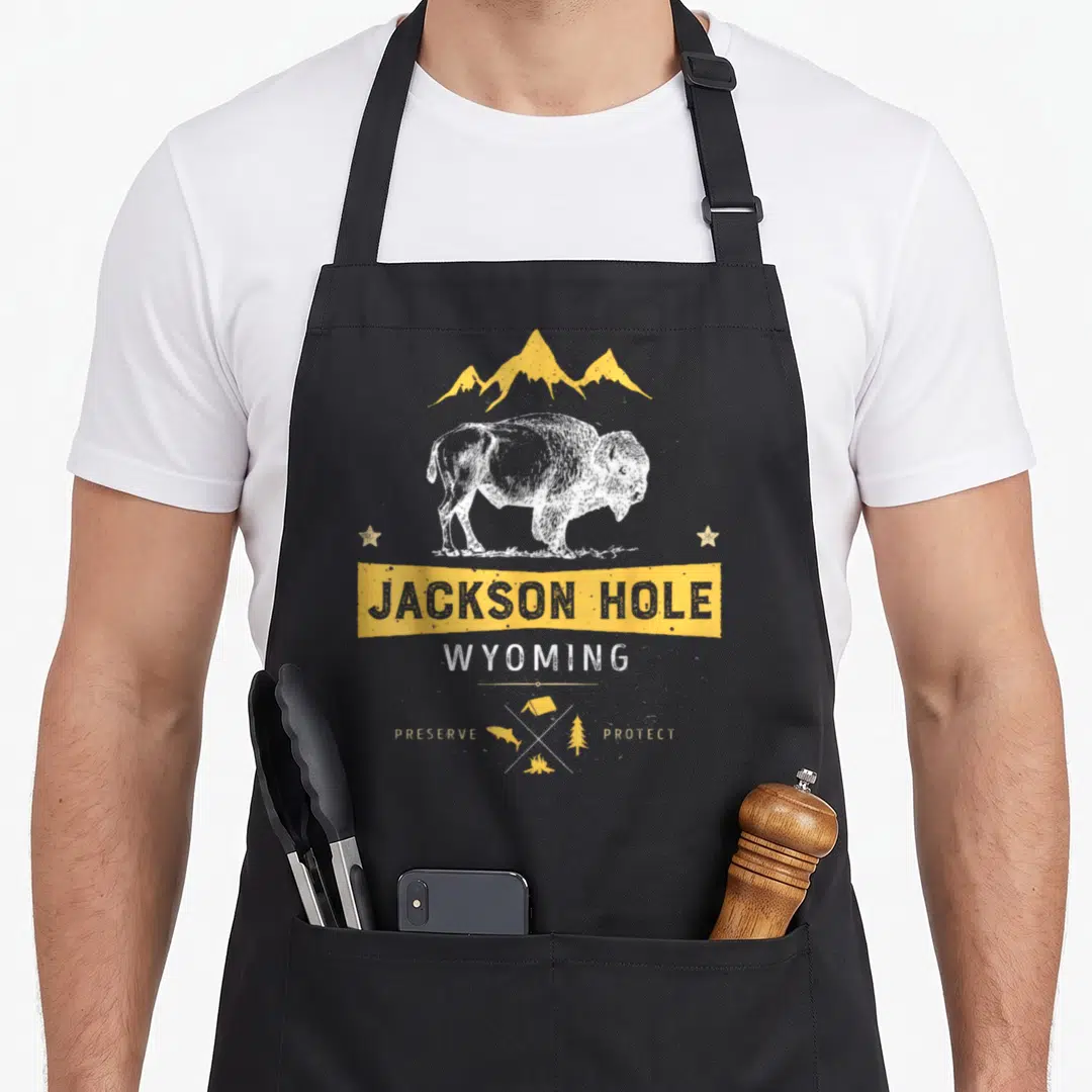Jackson buffalo apron - White bison under yellow mountains with Jackson Hole text and diamond camping icons — shown on a black cotton canvas apron against a white background