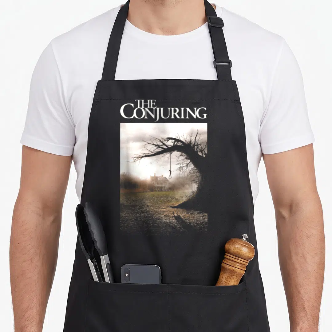 Conjuring paranormal investigation apron - White THE CONJURING text above photo of tree with noose and house — shown on a black cotton canvas apron against a white background