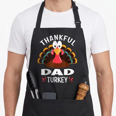Funny thankful thanksgiving apron - Cartoon turkey with arched THANKFUL text and stacked DAD TURKEY below flanked — shown on a black cotton canvas apron against a white background