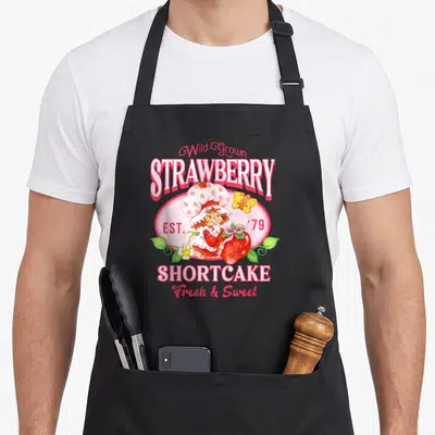 Shortcake strawberry apron - Cartoon girl holding red strawberry inside pink oval with Wild Grown and EST 79 text — shown on a black cotton canvas apron against a white background