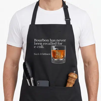 Bourbon e.a apron - White text reading Bourbon e-coli pun next to realistic amber bourbon glass on grey background — shown on a black cotton canvas apron against a white background