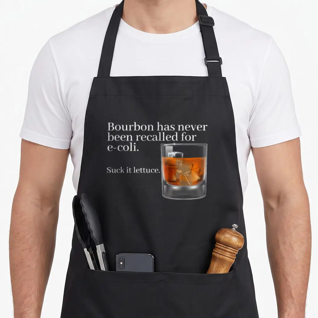 Bourbon e.a apron - White text reading Bourbon e-coli pun next to realistic amber bourbon glass on grey background — shown on a black cotton canvas apron against a white background
