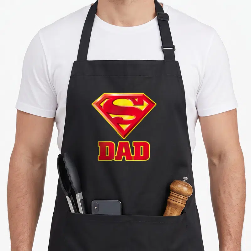Funny robin dad apron - Red and yellow shield logo above bold red DAD text on black fabric — shown on a black cotton canvas apron against a white background