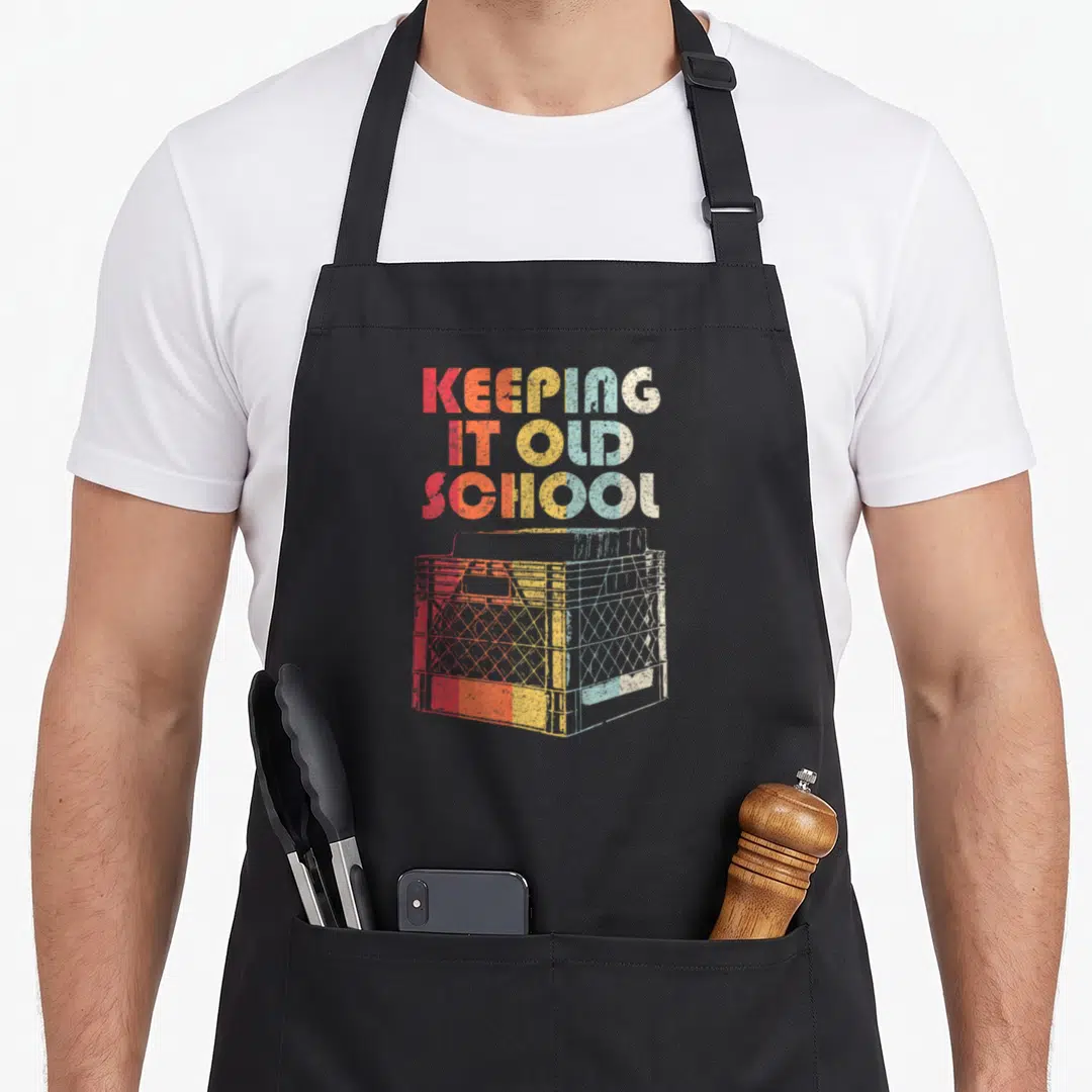 School vinyl apron - KEEPING IT OLD SCHOOL text and milk crate with red orange yellow blue grey gradient — shown on a black cotton canvas apron against a white background