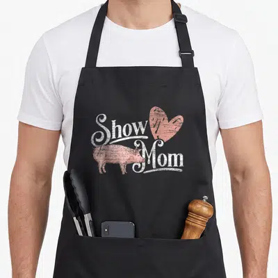 Show pig mom apron - Distressed white Show Mom text with pink pig and heart graphic — shown on a black cotton canvas apron against a white background