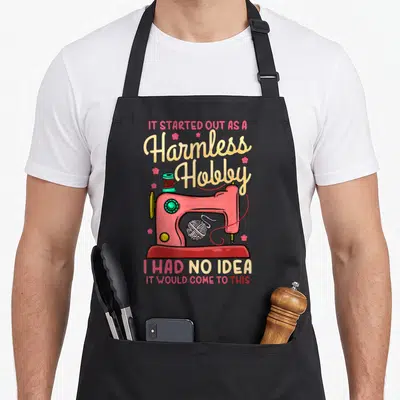 Sewing quilting apron - Pink cartoon sewing machine with thread spool yarn and quote IT STARTED OUT AS A Harmless Hobby — shown on a black cotton canvas apron against a white background
