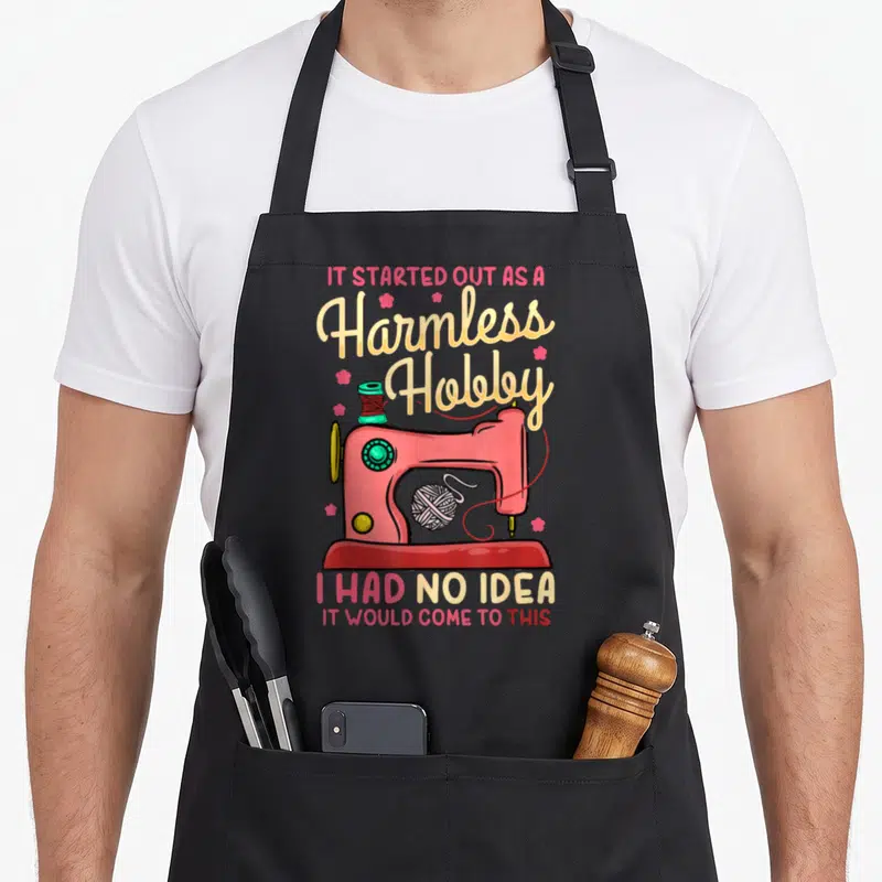 Sewing quilting apron - Pink cartoon sewing machine with thread spool yarn and quote IT STARTED OUT AS A Harmless Hobby — shown on a black cotton canvas apron against a white background