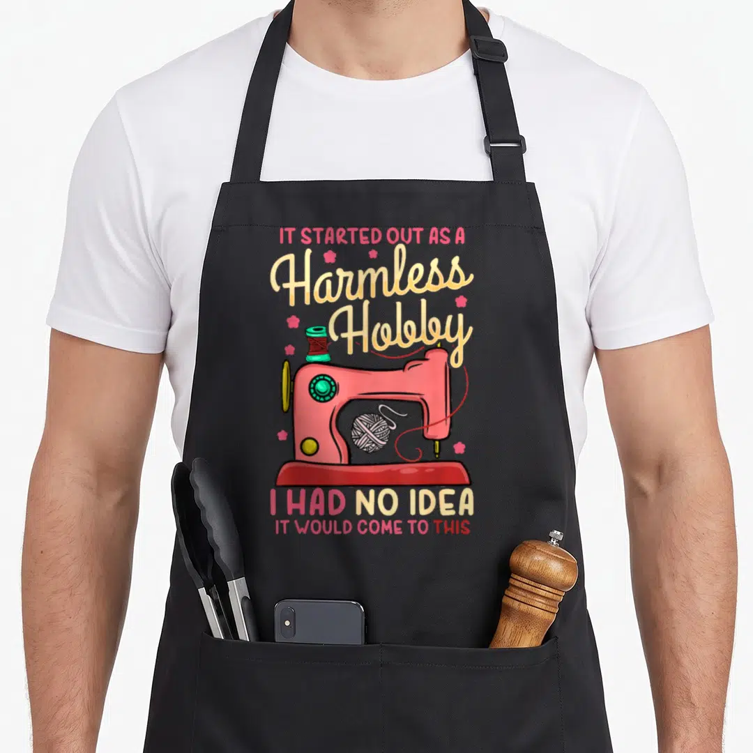Sewing quilting apron - Pink cartoon sewing machine with thread spool yarn and quote IT STARTED OUT AS A Harmless Hobby — shown on a black cotton canvas apron against a white background