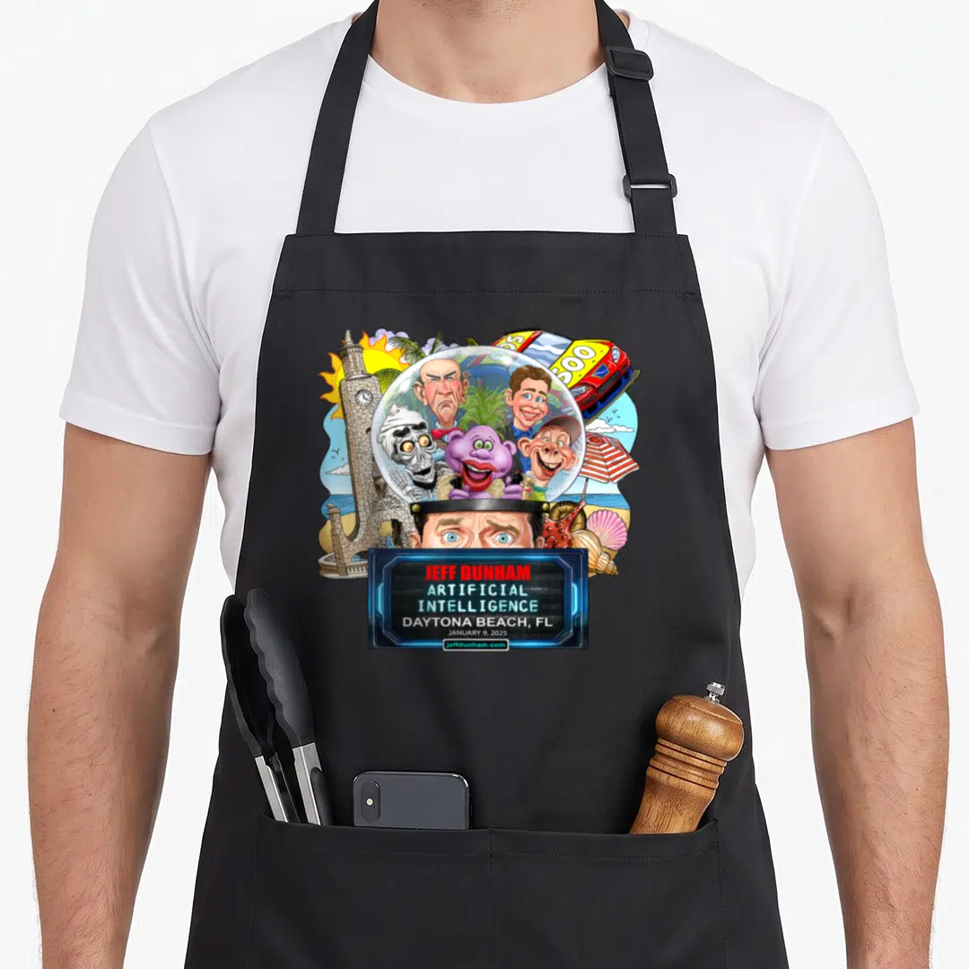 Jeff daytona apron - Jeff Dunham cartoon head with crystal ball, four puppets, clock tower, race car, and event text — shown on a black cotton canvas apron against a white background