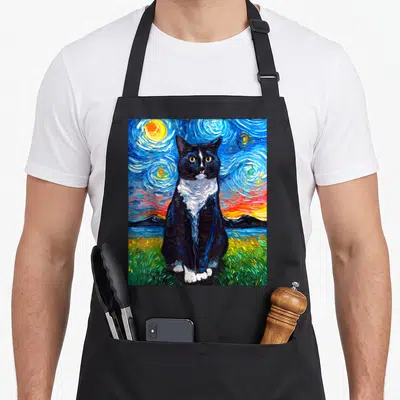 Cat kitty tuxedo apron - Realistic black and white cat sitting in green grass before blue swirling sky with sun — shown on a black cotton canvas apron against a white background