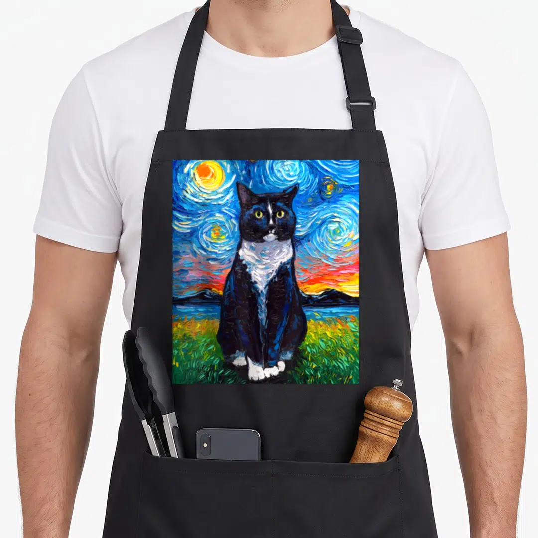 Cat kitty tuxedo apron - Realistic black and white cat sitting in green grass before blue swirling sky with sun — shown on a black cotton canvas apron against a white background