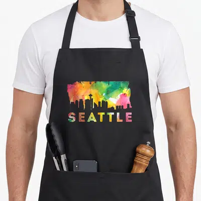 Skyline watercolor seattle apron - Colorful watercolor skyline silhouette above SEATTLE text in red yellow green blue — shown on a black cotton canvas apron against a white background