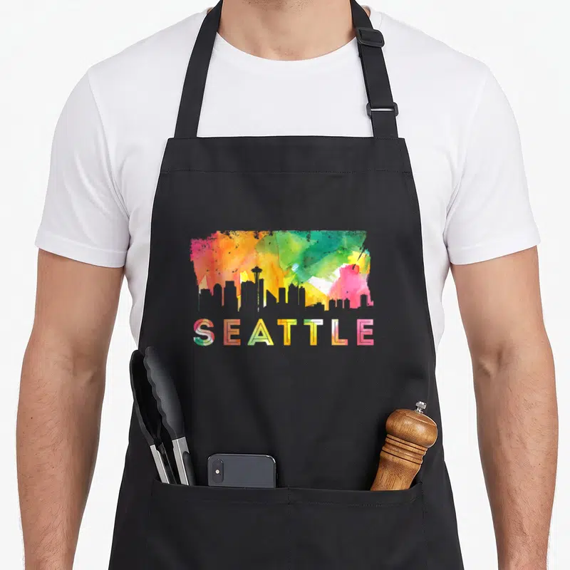 Skyline watercolor seattle apron - Colorful watercolor skyline silhouette above SEATTLE text in red yellow green blue — shown on a black cotton canvas apron against a white background