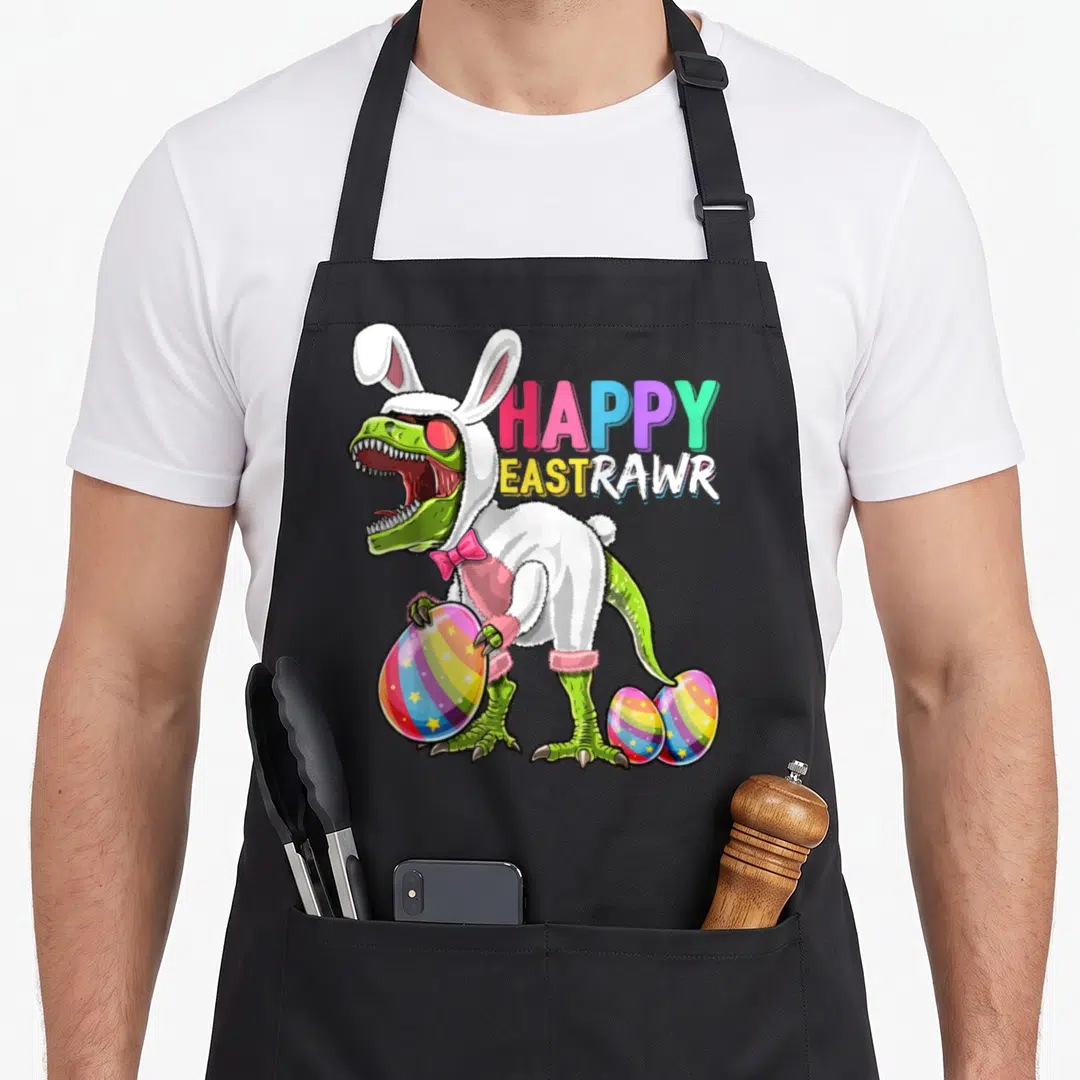 Eastrawr easter egg apron - Green T-Rex in white bunny suit holding striped rainbow egg next to HAPPY EASTRAWR text — shown on a black cotton canvas apron against a white background
