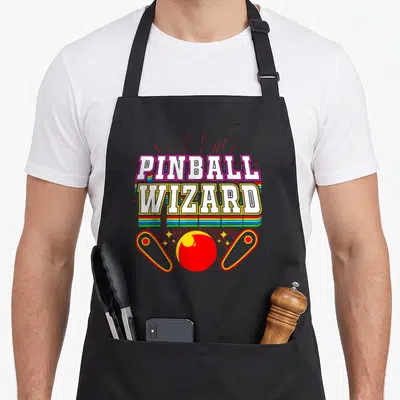 Wizard arcade apron - Stacked PINBALL WIZARD text over rainbow stripes with red pinball ball and two flippers — shown on a black cotton canvas apron against a white background