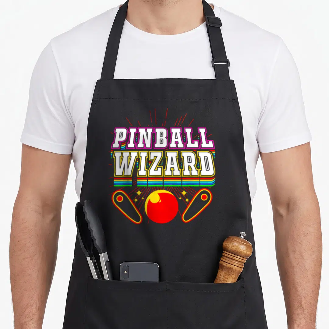 Wizard arcade apron - Stacked PINBALL WIZARD text over rainbow stripes with red pinball ball and two flippers — shown on a black cotton canvas apron against a white background