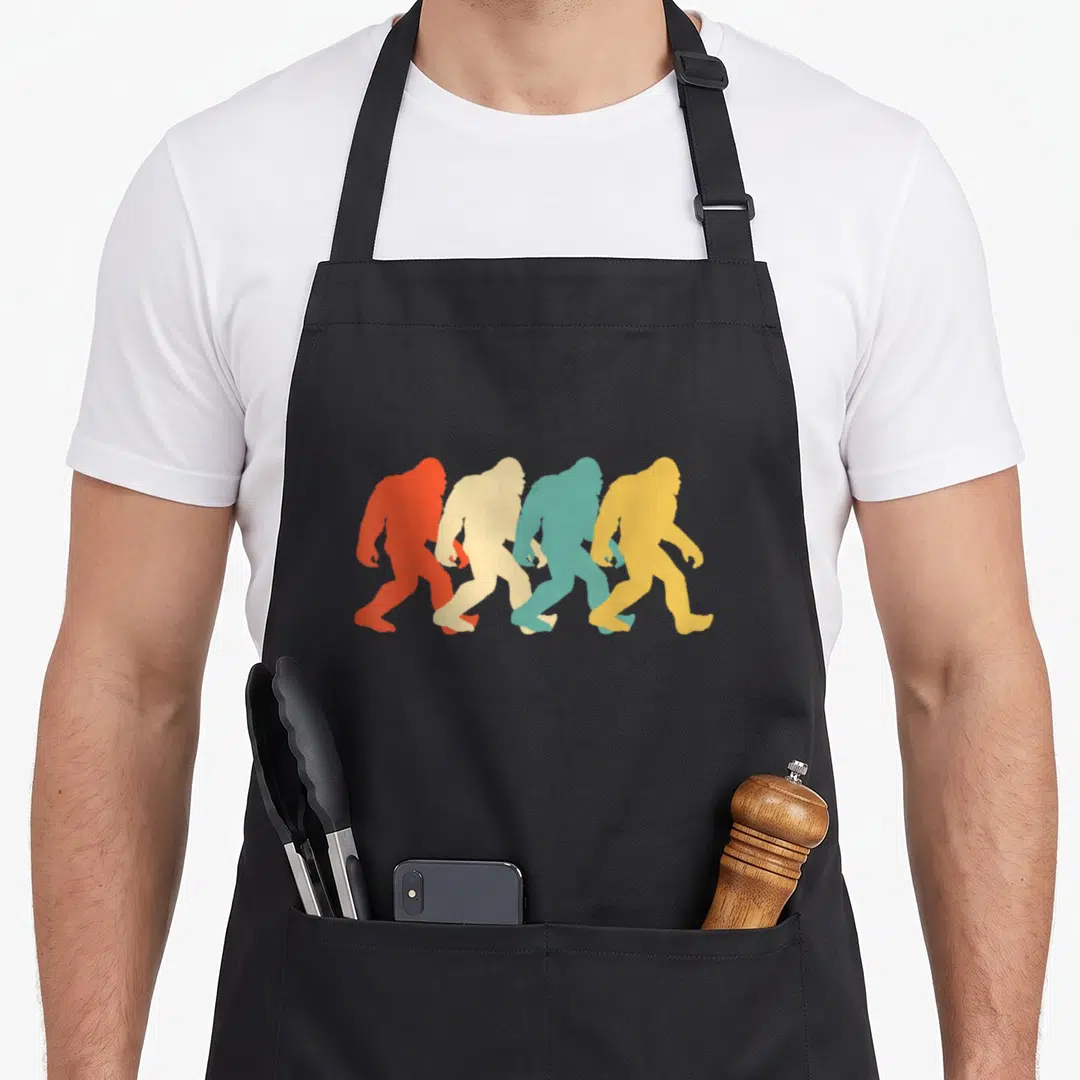Pop bigfoot apron - Four colorful Bigfoot silhouettes walking right in red beige teal yellow — shown on a black cotton canvas apron against a white background