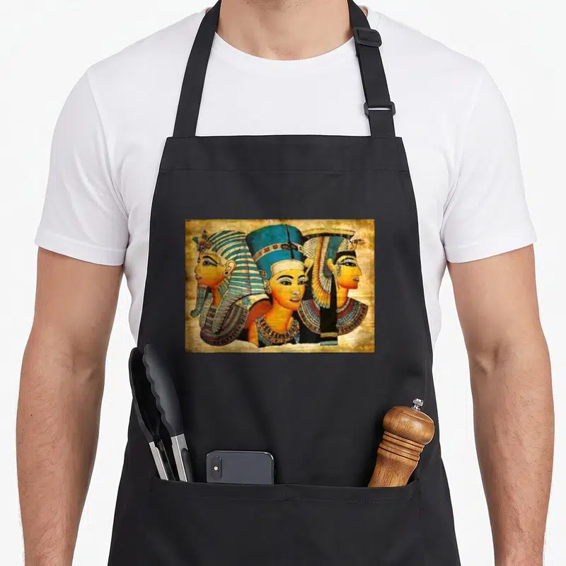 Cleopatra egyptian apron - Three overlapping Egyptian royal figures in blue, gold, orange, and brown on textured brown — shown on a black cotton canvas apron against a white background