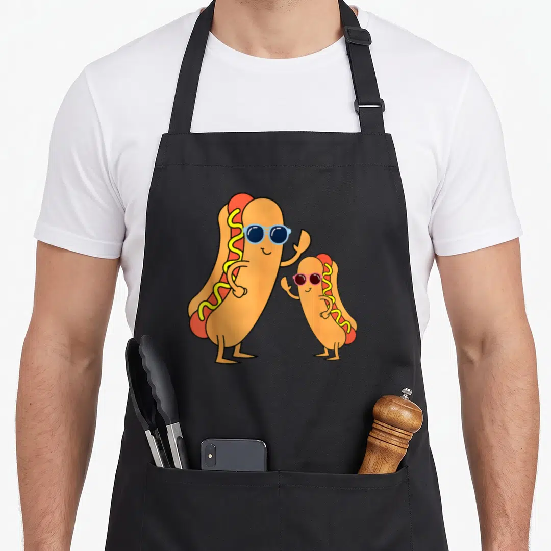 Frank sausage apron - Two cartoon hot dogs wearing sunglasses and waving, colored in orange, red, yellow, and blue — shown on a black cotton canvas apron against a white background