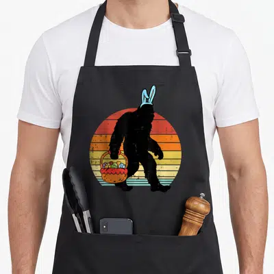 Bigfoot easter egg apron - Black Bigfoot silhouette with blue bunny ears holding an orange basket of eggs against — shown on a black cotton canvas apron against a white background