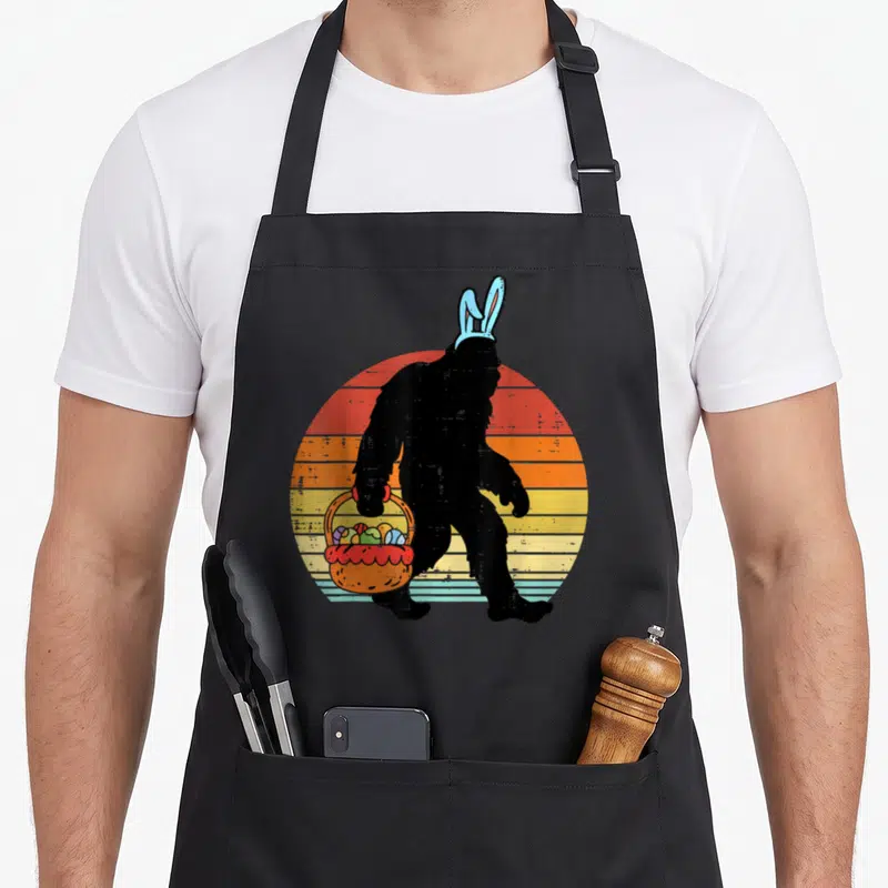 Bigfoot easter egg apron - Black Bigfoot silhouette with blue bunny ears holding an orange basket of eggs against — shown on a black cotton canvas apron against a white background