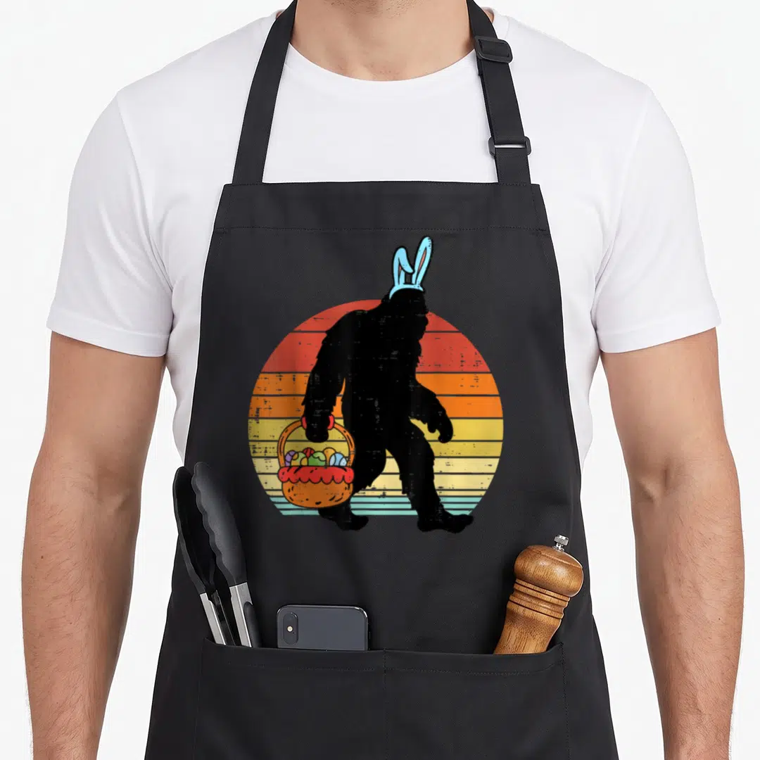 Bigfoot easter egg apron - Black Bigfoot silhouette with blue bunny ears holding an orange basket of eggs against — shown on a black cotton canvas apron against a white background