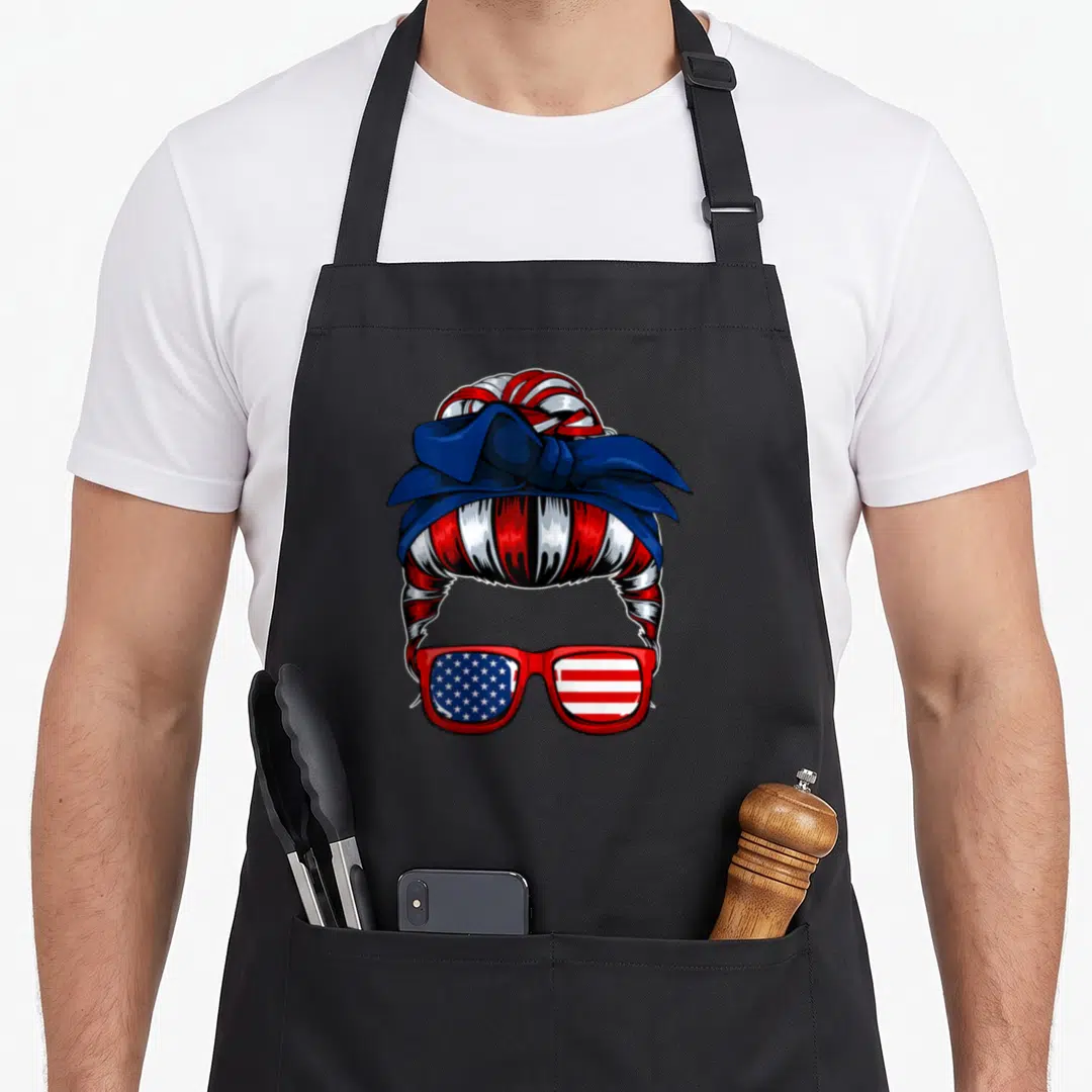 July messy aprons - Cartoon messy bun with red white blue stripes and patriotic sunglasses on black apron — shown on a black cotton canvas apron against a white background