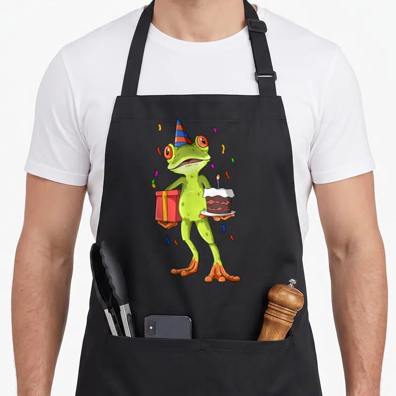 Birthday frog aprons - Green cartoon frog holding red gift and cake with party hat and confetti — shown on a black cotton canvas apron against a white background