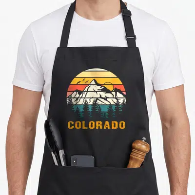 Hiker pride apron - Retro circular sunset with mountains, birds, trees, and COLORADO text in orange, teal, white — shown on a black cotton canvas apron against a white background