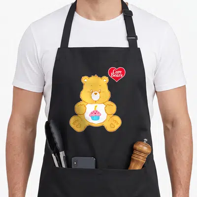 Cool birthday apron for hosting - Yellow bear holding pink cupcake with candle and red Care Bears heart logo — shown on a black cotton canvas apron against a white background