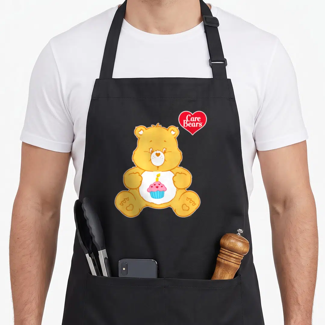 Cool birthday apron for hosting - Yellow bear holding pink cupcake with candle and red Care Bears heart logo — shown on a black cotton canvas apron against a white background