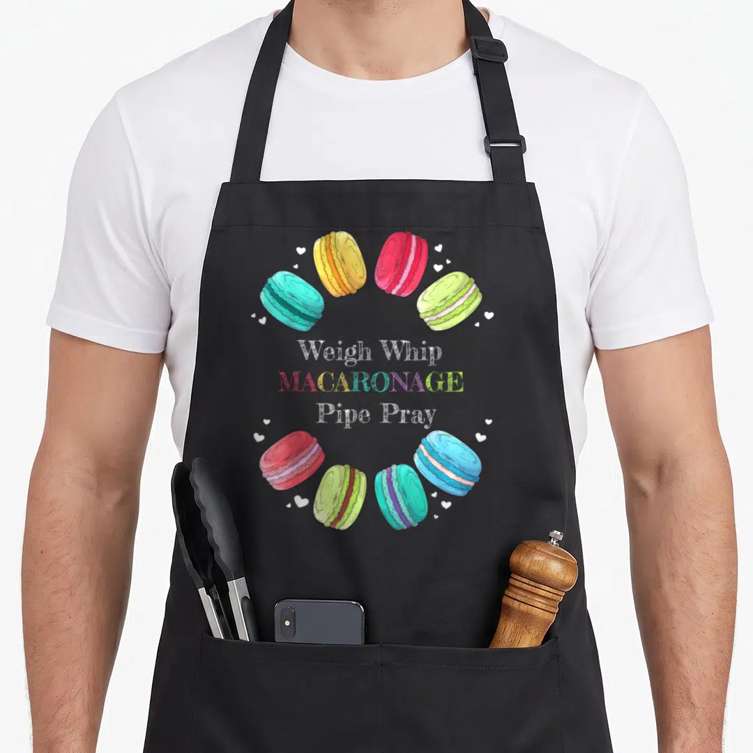 Weigh macaron apron - Eight colorful macarons and hearts circling Weigh Whip Macaronage Pipe Pray text — shown on a black cotton canvas apron against a white background