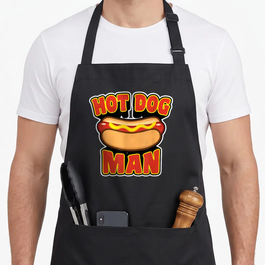 Retro hot dog apron - Red and yellow HOT DOG MAN text framing a cartoon sausage with mustard on a bun — shown on a black cotton canvas apron against a white background