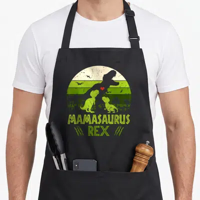 Mamasaurus dinosaur apron - Retro T-Rex silhouette with two baby dinos, red heart, and MAMASAURUS REX text in lime — shown on a black cotton canvas apron against a white background