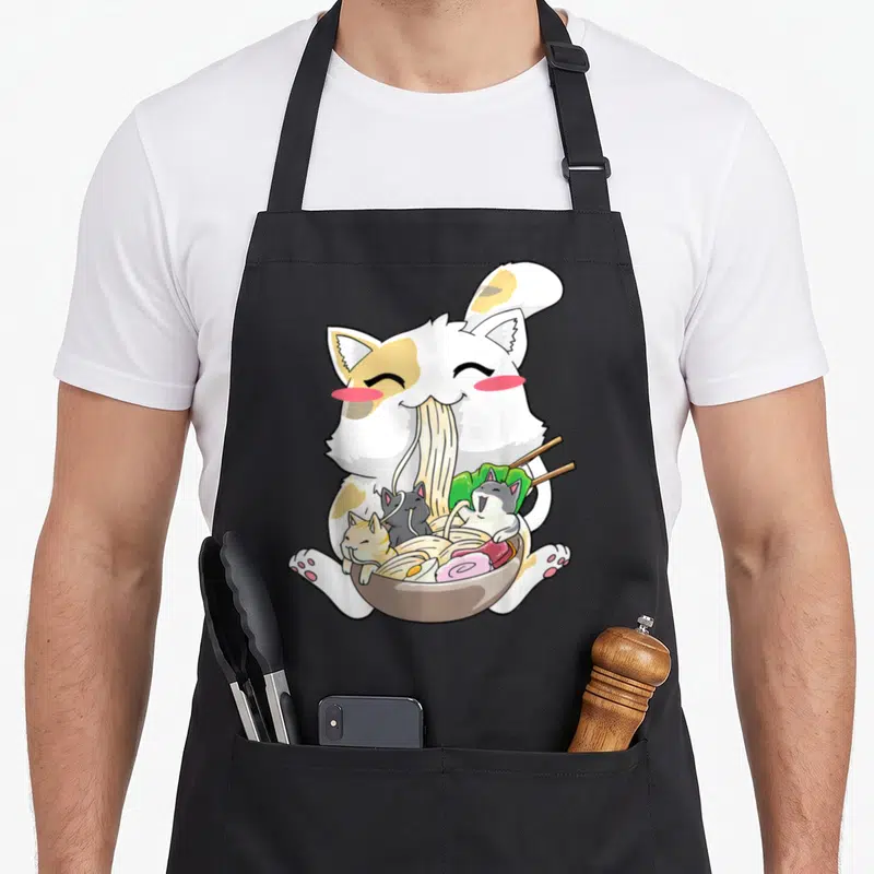 Japanese kawaii cooking apron - Large white and orange cat eating ramen with three grey kittens inside a brown bowl — shown on a black cotton canvas apron against a white background