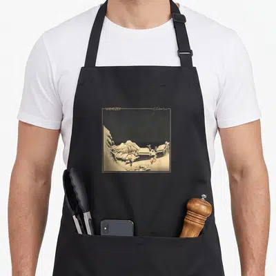 Weezer caponata apron - Square snowy village print with weezers and Pinkerton text on beige border — shown on a black cotton canvas apron against a white background