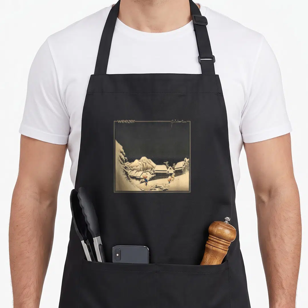 Weezer caponata apron - Square snowy village print with weezers and Pinkerton text on beige border — shown on a black cotton canvas apron against a white background