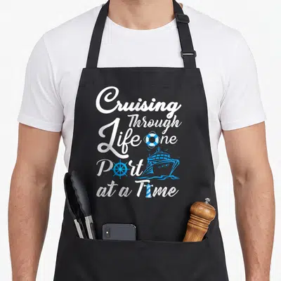 Through cruising apron - Cruising Through Life one Port at a Time text with blue ship, lifebuoy, wheel, and lighthouse — shown on a black cotton canvas apron against a white background