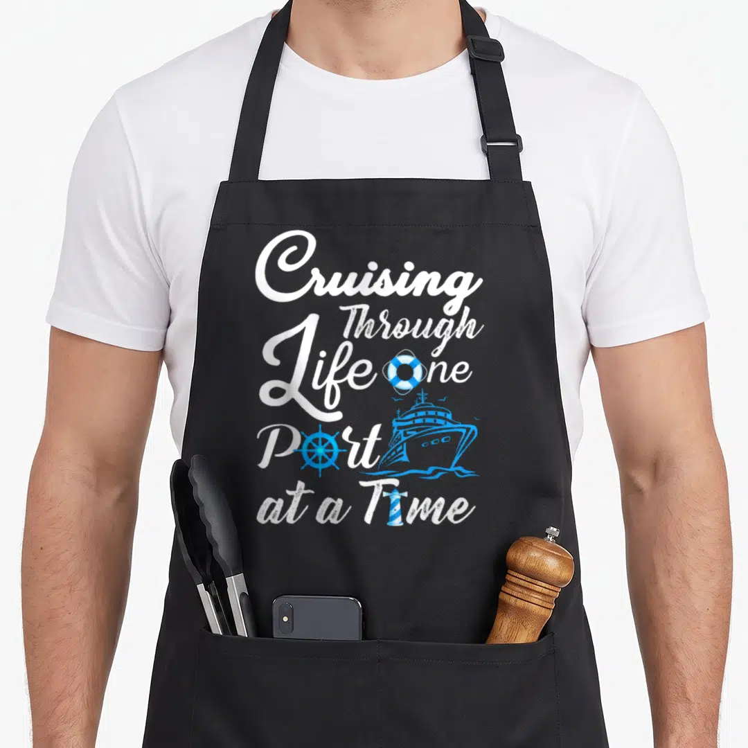 Through cruising apron - Cruising Through Life one Port at a Time text with blue ship, lifebuoy, wheel, and lighthouse — shown on a black cotton canvas apron against a white background
