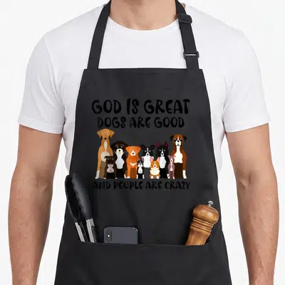 Retro god cooking with dog apron - Cartoon dogs in black, brown, white, and orange between text blocks reading God is — shown on a black cotton canvas apron against a white background