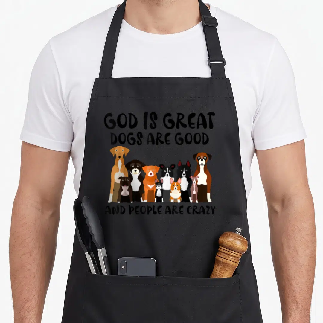 Retro god cooking with dog apron - Cartoon dogs in black, brown, white, and orange between text blocks reading God is — shown on a black cotton canvas apron against a white background