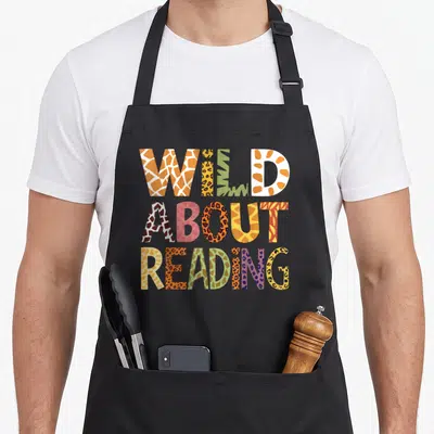 Animal teacher apron - Wild About Reading text filled with giraffe, leopard, and zebra patterns in orange and purple — shown on a black cotton canvas apron against a white background