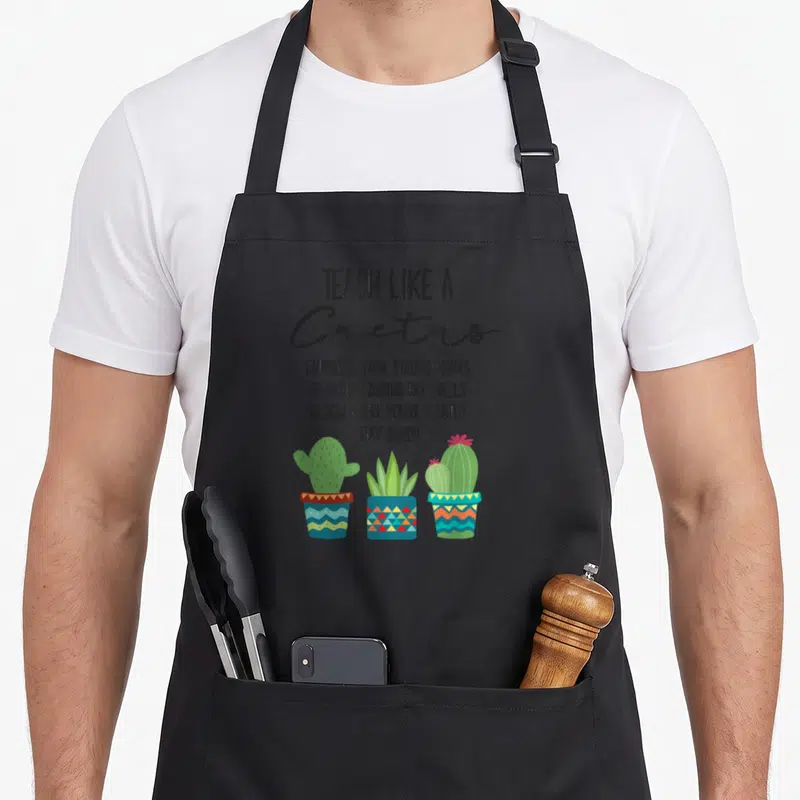 Teach cactus teacher apron - Stacked text TEACH LIKE A CACTUS above three potted cacti in green blue and pink cartoon — shown on a black cotton canvas apron against a white background