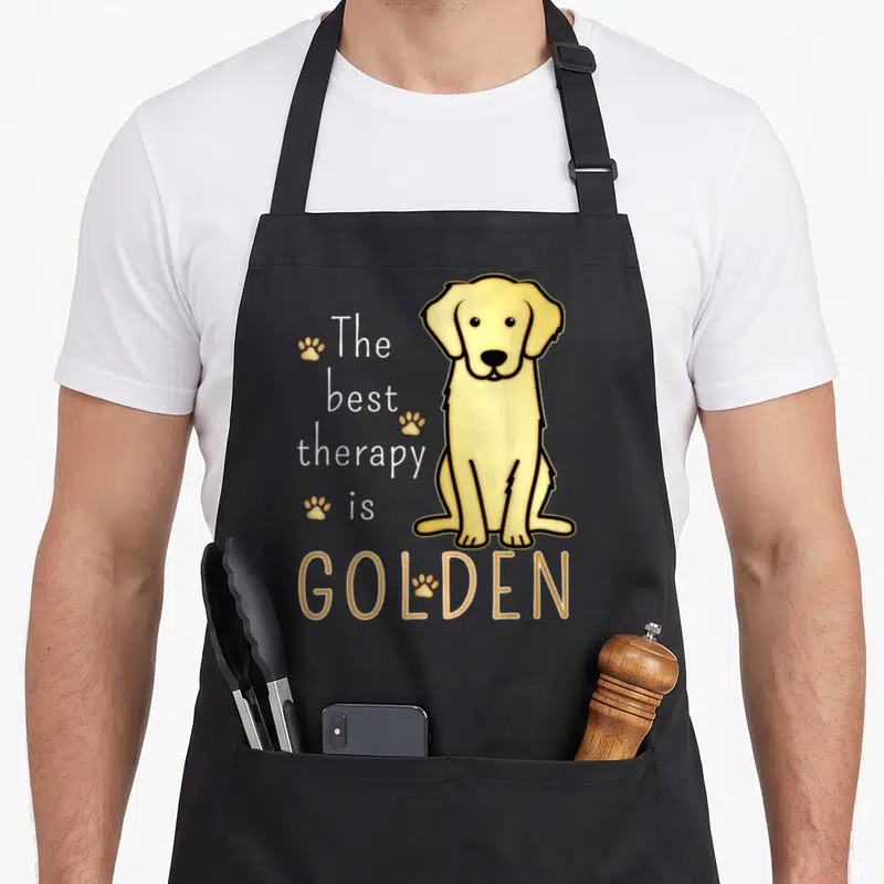 Therapy golden retriever apron - Cartoon golden dog sitting next to stacked text The best therapy is GOLDEN — shown on a black cotton canvas apron against a white background