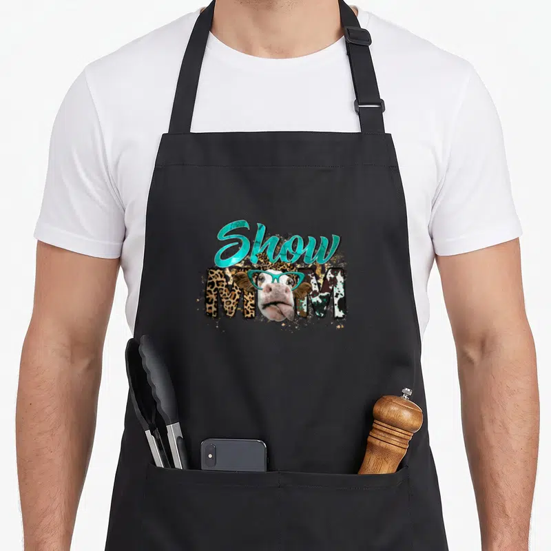 Cowhide mother's day aprons - Cartoon cow head with teal glasses overlapping leopard print Show MOM text — shown on a black cotton canvas apron against a white background